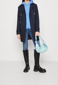 Abrigo azul marino de doble botonadura, sudadera con capucha azul clara, bolso azul, jeans azules y botas negras hasta la rodilla con suelas gruesas.