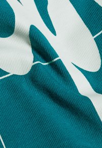 Teal stof med en struktureret overflade præsenterer store hvide grafiske elementer. Designet er dristigt og abstrakt, hvilket skaber visuel kontrast.