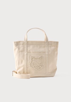 Sac fourre-tout en toile beige avec deux anses, une bandoulière amovible et une tête de loup brodée sur la poche avant.