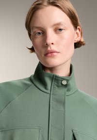 Giacca verde con colletto alto realizzata in tessuto testurizzato, dotata di chiusura a pressione e due tasche frontali, con un design pulito e moderno.