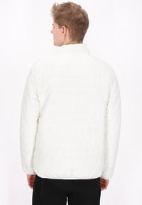 Geacă puffer albă cu textură matlasată, guler în stand, și mâneci lungi, purtată de o persoană cu spatele, având un design simplu și un aspect ușor.