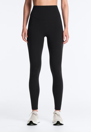 Vue de face d'une personne portant des leggings noirs taille haute et des chaussures de sport blanches, debout devant un fond blanc uni.