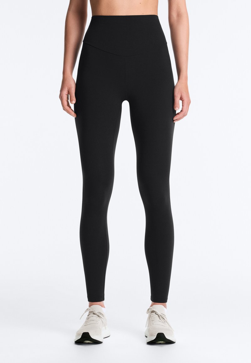 Elölnézet egy személyről, aki magas derekú fekete leggingset és fehér sportcipőt visel, egy sima fehér háttér előtt állva.