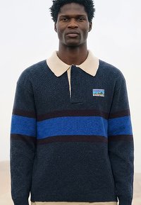 Uomo che indossa un maglione blu navy a maniche lunghe con una larga striscia blu e colletto beige, in piedi contro uno sfondo chiaro e semplice.