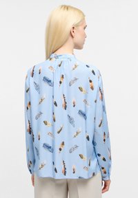 Blusa azzurra chiara con stampa di piume in vari colori. Il design presenta un colletto alla cinese e maniche lunghe con polsini abbottonati.