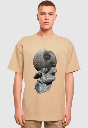 Beige bomulds t-shirt med et grafisk motiv af en skulptureret figur, der holder en tekstureret fodbold med "FCH" præget. Afslappet pasform, korte ærmer.