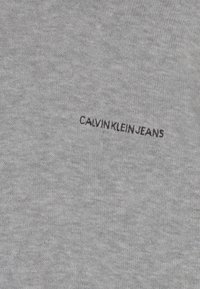 Szara bawełniana tkanina o gładkiej fakturze, z haftem "CALVIN KLEIN JEANS" w czerni wzdłuż prawej strony.