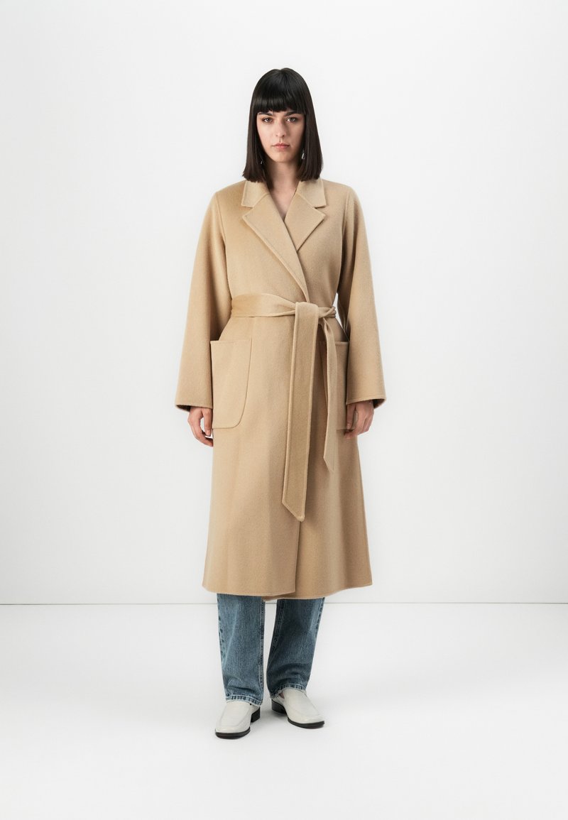 Kvinna med rakt mörkt hår iförd en lång beige kåpa med bälte, blå jeans och vita skor, stående mot en vit bakgrund.