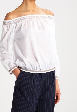 Femme portant un chemisier blanc à épaules dénudées avec un bord élastique, associé à un pantalon bleu marine à fines rayures blanches, main dans la poche.