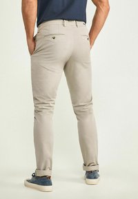 Beige slim-fit pants with a smooth texture, featuring two back pockets and decorative stitching, paired with blue sneakers.