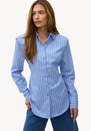 Femme aux cheveux longs portant une chemise boutonnée à rayures verticales bleues et blanches et un jean bleu, debout avec une main dans la poche.