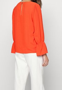 Blouse rouge texturée avec manches bouffantes et détail dos en forme de goutte, associée à un pantalon blanc. Le tissu semble léger et fluide.