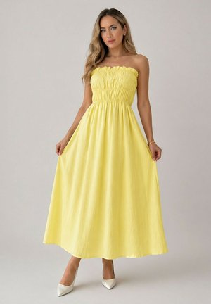 Femme aux longs cheveux portant une robe maxi jaune sans bretelles et froncée, avec des talons blancs, debout devant un fond clair uni.