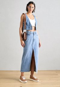 Femme portant des lunettes transparentes, un crop top blanc, un gilet en jean sans manches, une jupe en jean taille haute fendue à l'avant, des baskets marron et un sac à bandoulière marron.