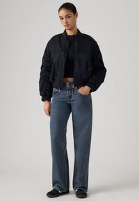 Μαύρο μπουφάν bomber με ελαστικά μανίκια, φορεμένο πάνω από εφαρμοστή μαύρη μπλούζα. Συνδυάζεται με φαρδιά μπλε τζιν και μαύρα παπούτσια. Ομαλή υφή υφάσματος.