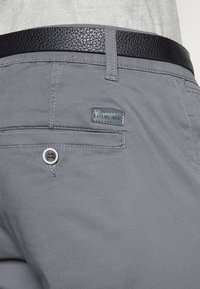 Pantalones grises de tela suave, con un cierre de botón, costuras de acento y una etiqueta rectangular de la marca. Cinturón negro texturizado arriba.