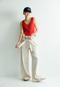 Rood geribbeld tanktop, beige wijde broek met strik, en lichte schoenen. Een zwarte pet met witte tekst maakt de outfit af tegen een neutrale achtergrond.