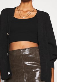 Cardigan noir en maille avec des boutons en accent, porté sur un haut court noir. Une jupe verte olive brillante et texturée complète la tenue.