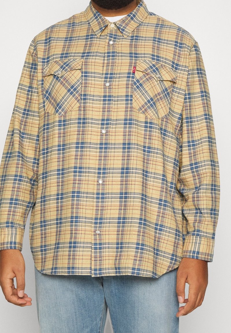 Personne portant une chemise à carreaux beige, bleue et rouge avec boutons, avec deux poches sur la poitrine et un jean bleu clair.