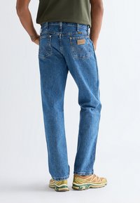 Person iført blå Wrangler straight-leg jeans og gule Salomon trail-løbesko, stående med ryggen til en ensfarvet baggrund.