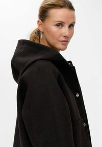 Cappotto nero con cappuccio, realizzato in tessuto morbido, dotato di bottoni a pressione argentati e tasche laterali. Il design presenta una vestibilità morbida e un cappuccio ben visibile.