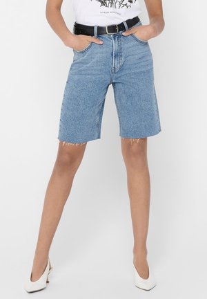 Person iført lyseblå cowboy-shorts til knæene med frynser, sort bælte, hvide hæle og en hvid t-shirt med tryk.