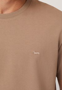 Tan crew-neck t-shirt i mjukt tyg, med en liten broderad taxdesign på vänster bröst.