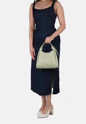 Femme en robe sans manches bleu foncé tenant un sac à main vert clair, portant des chaussures blanches à talons carrés et un bracelet argenté, debout devant un fond blanc.