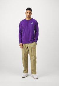 Maglietta viola a maniche lunghe con logo Nike verde; pantaloni khaki con loghi NBA e Nike; sneakers bianche; tessuto morbido, vestibilità rilassata.
