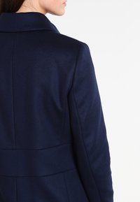 Manteau sur mesure bleu marine avec un col structuré, tissu lisse, couture cintrée à la taille et sans fermetures visibles ; design épuré et minimaliste.