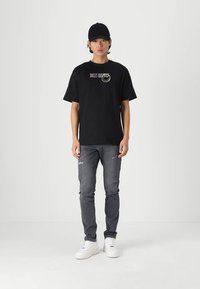 Camiseta de algodón negra con gráfico "Smiley Objects" en blanco y amarillo, combinada con jeans grises desgastados y zapatillas blancas. Hombre de pie en un entorno neutro.