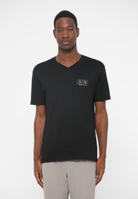 Άνδρας φορώντας μαύρη μπλούζα V-neck Armani Exchange με λογότυπο στο στήθος και ανοιχτό γκρι παντελόνι μπροστά σε λευκό φόντο.