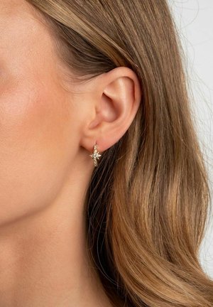 Nahaufnahme eines Ohrs mit hellbraunem Haar, das einen kleinen goldenen Creolenohrring mit einem sternförmigen Kristallanhänger trägt.