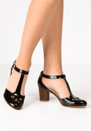 Beine, die schwarze T-Riemchen-Pumps aus Lackleder mit Cut-out-Details und einem Holzblockabsatz vor weißem Hintergrund tragen.
