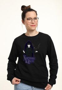 Mujer con gafas y un moño que lleva una sudadera negra con un personaje gótico que tiene un cuervo y el texto "Sombras Nocturnas y Cuervos".