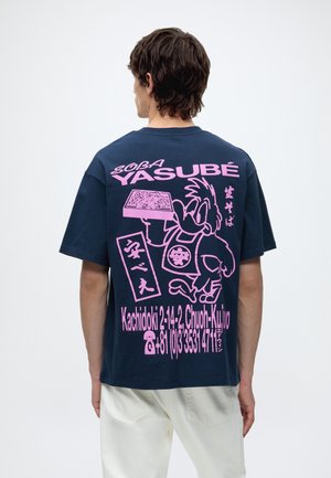 Mand med ryggen mod, iført navyblå t-shirt med stort pink tegneserie- og japansk tekst, der promoverer en soba-restaurant og kontaktoplysninger.