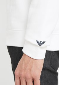 White sweatshirt with ribbed cuffs and a textured black logo on the left sleeve. Soft fabric, fitted shape, and minimalistic design.