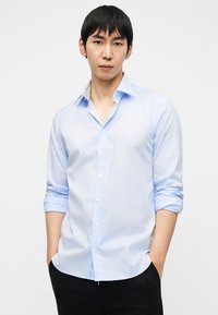 Ανοιχτό μπλε πουκάμισο με κουμπιά, με διασπειρόμενο γιακά, γυρισμένα μανίκια και λεία υφή. Συνδυασμένο με σκούρο παντελόνι, δημιουργώντας αντίθεση.