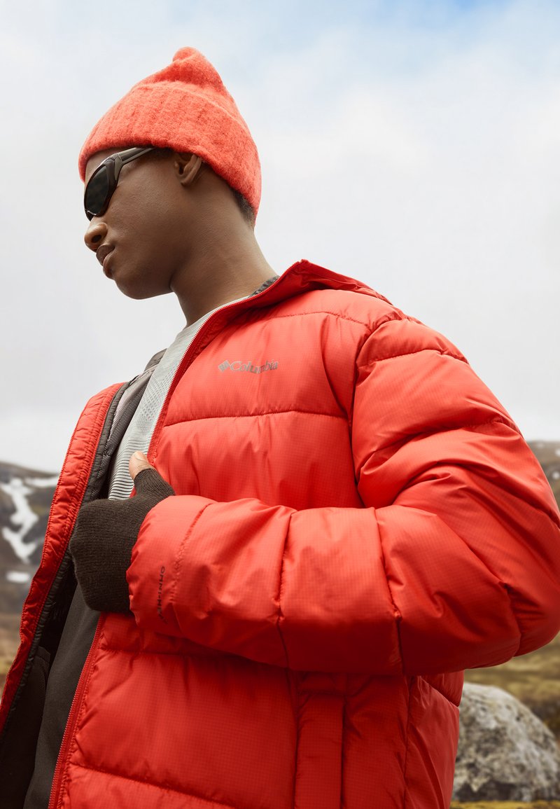 Chaqueta de plumas roja con un diseño acolchado, que incluye una capucha y un detalle de logotipo. Combinada con un gorro de punto marrón y unas gafas de sol oscuras.