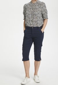 Marinblå knälånga cargoshorts med sidofickor, kombinerade med en kortärmad mönstrad blus i ljusa färger och vita sneakers.