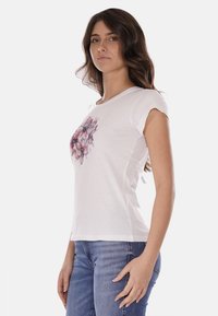 T-shirt bianca con motivo floreale in tonalità rosa, maniche corte, tessuto leggero e design aderente. Indossata con jeans blu.