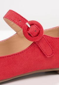 Chaussure en daim rouge avec un bout arrondi, dotée d'une sangle maintenue par une boucle circulaire et de légères coutures sur le dessus.
