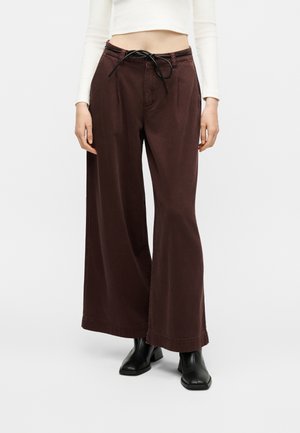 Femme portant un pantalon marron taille haute à jambes larges avec cordon noir, un haut blanc court à manches longues et des bottines noires.