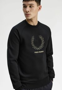 Felpa nera con scollo rotondo, polsini a coste e una corona di alloro ricamata in oro con il testo "FRED PERRY" al centro.