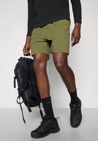 Person wearing olive-green shorts, black long-sleeve shirt, black hiking shoes, black socks, holding a black backpack by the handle.