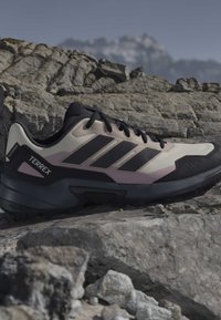 Παπούτσι για ορεινό τρέξιμο με μαύρες ρίγες και μπεζ επάνω μέρος, που βρίσκεται σε βραχώδες ορεινό έδαφος με θολές κορυφές στο φόντο.