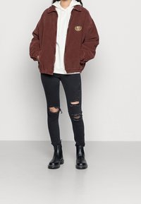 Dunkelrote Cordjacke mit Kragen, versehen mit einem gestickten Emblem. Kombiniert mit einem weißen Hoodie, ausgewaschenen schwarzen Jeans und schwarzen Stiefeln.