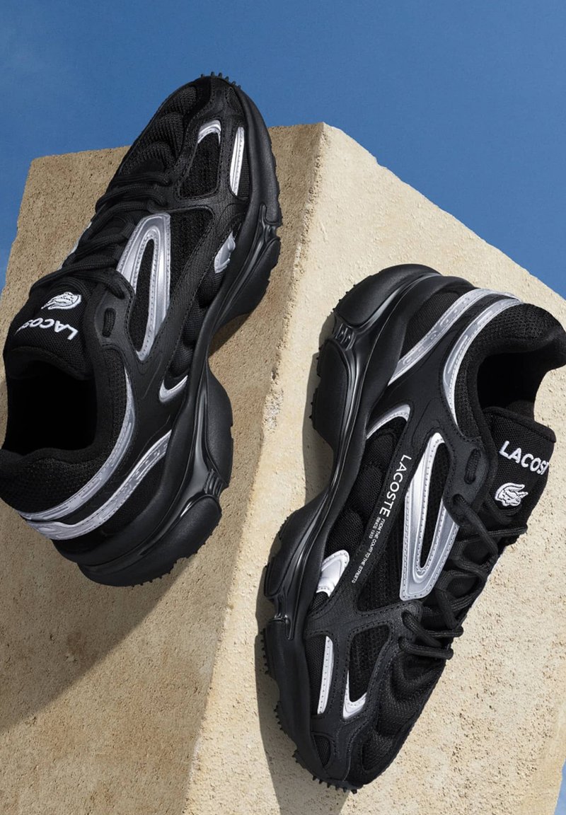 Zapatillas deportivas negras con acentos plateados y parte superior de malla texturizada. Presenta una suela gruesa y un diseño de cordones visible, de la marca Lacoste.