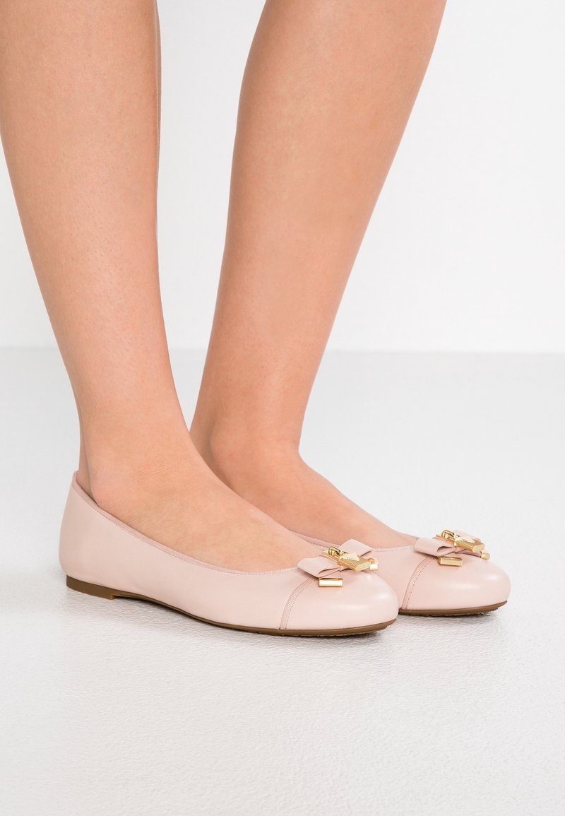 Ballerines en cuir nude avec un bout arrondi et un nœud orné de détails dorés, présentées sur un fond blanc lisse.