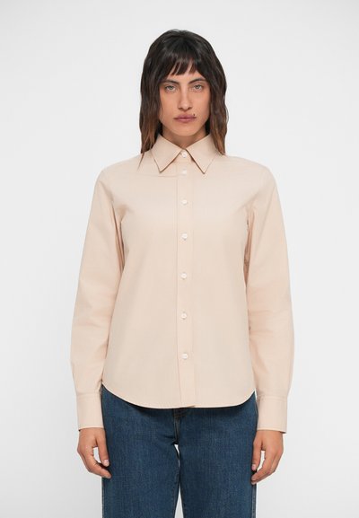 Camisa button-up beige con cuello puntiagudo, mangas largas y textura suave. Presenta un dobladillo recto y se combina con jeans oscuros.